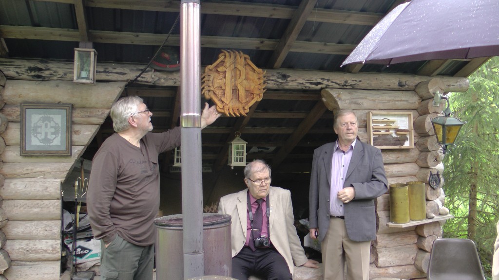 Päivänsankari esittelee isoa puusta veistettyä Rukajärven R-merkkiä. Istumassa Timo Laukkanen ja hänen vieressään Raimo Jokisalmi. - Kuva: Antero Huttunen.